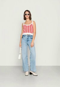 Top curto de malha xadrez vermelho e branco, calças largas jeans azul claro, botas ankle bege, pequena mala branca e óculos de sol redondos pretos.