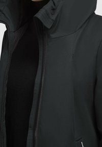 Chaqueta oscura, mate, con cremallera y capucha, que presenta una textura suave, detalles cosidos y bolsillos laterales discretos.