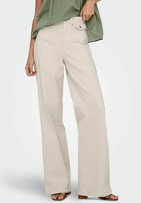 Pantalons larges beige en coton doux, avec des poches avant, une texture subtile et une taille haute. Associés à un haut vert.