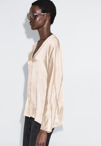 Beige, texturovaný top s dlouhým rukávem a výstřihem do V, doplněný černými džíny a oversize slunečními brýlemi, umístěný na neutrálním pozadí.