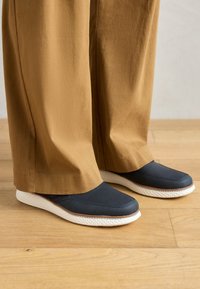 Baskets navy à enfiler avec dessus texturé, semelle en caoutchouc blanche et accents marron. Portées avec un pantalon ample beige sur un sol en bois.