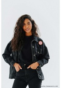 Czarna kurtka dżinsowa z zapięciem na guziki i kieszeniami, noszona na czarnej koszuli, zestawiona z wysokim stanem czarnych dżinsów. Posiada okrągłą pomarańczową naszywkę.