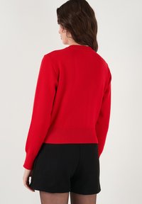 Roter Strickpullover mit langen Ärmeln, geripptem Kragen und Bündchen, cropped Design, kombiniert mit schwarzen Shorts. Glatte Textur und figurbetonte Form.