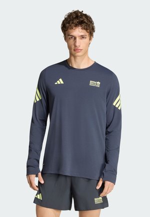 Ung mand iført navyblå langærmet skjorte og shorts med neon Adidas-logoer og teksten "BMW Berlin Marathon", står mod en ensfarvet baggrund.