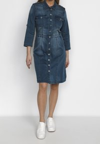 Jeanimekko tummansinisestä denimistä, pitkät hihat, kaksi rintataskua, nappisuljenta edessä ja vyötäröä korostava malli, päättyy juuri polven ylle.