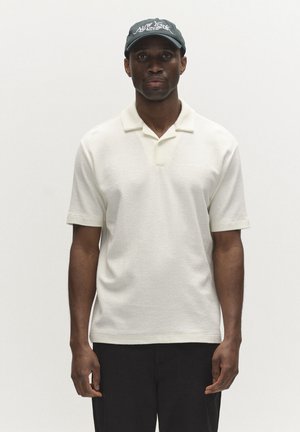 Mężczyzna ubrany w biały polo z krótkim rękawem, czarne spodnie i szarą czapkę z daszkiem New York, stojący na jednolitym jasnym tle.