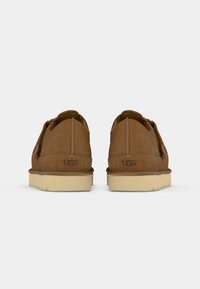 Bruine suède schoenen met een tan rubberen loopzool, met een ronde vorm, gestikte details en een UGG-logo op de hiel.