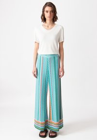 Wijduitlopende turquoise broek met kleurrijke zigzagpatronen en een decoratieve zoom. Gecombineerd met een aansluitend wit V-hals top.
