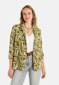 Blazer vert avec un motif de feuilles blanches ; manches trois-quarts et design ouvert à l'avant. Associé à un top blanc et un jean gris clair.