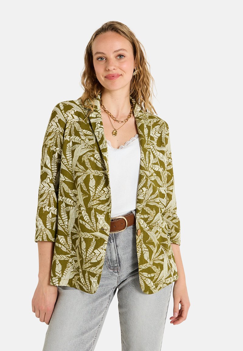 Blazer vert avec un motif de feuilles blanches ; manches trois-quarts et design ouvert à l'avant. Associé à un top blanc et un jean gris clair.