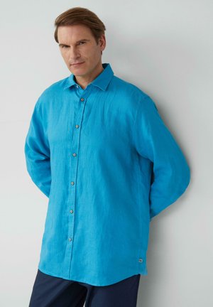 Chemise en lin bleu clair à boutons, avec un col, des manches longues et un ourlet arrondi, présentant des boutons marron et une petite étiquette logo sur le côté.
