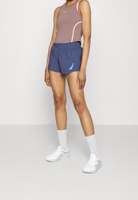 Mujer con una camiseta sin mangas morada, pantalones cortos deportivos azules con el logo de Nike, calcetines blancos y zapatillas Nike blancas de pie sobre un suelo gris.