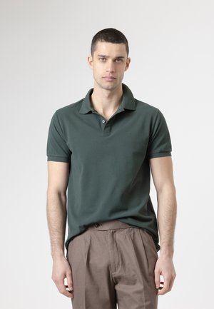 Joven de pie, de postura recta, vistiendo una camiseta tipo polo verde oscuro y pantalones marrón claro, frente a un fondo claro y liso.