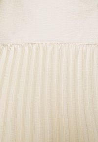 Crèmekleurige stof met een gestructureerde bovenhelft en gladde plooien in de onderhelft, wat verticale lijnen creëert. Zacht, lichtgewicht materiaal.