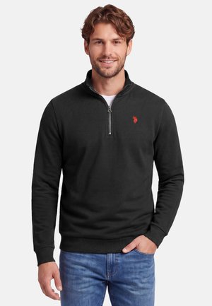 Lächelnder Mann, der einen schwarzen Viertel-Reißverschluss-Pullover mit kleinem rotem Logo auf der Brust und blaue Jeans trägt, Hand in der Tasche vor weißem Hintergrund.