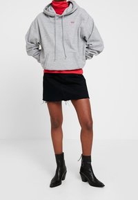Sweat-shirt gris à capuche avec cordons, porté par-dessus un col roulé rouge, associé à une jupe en jean noire et des bottines.