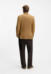 Blazer beige con due ventagli posteriori, spalle strutturate e una texture liscia, abbinato a pantaloni marroni scuri, larghi.