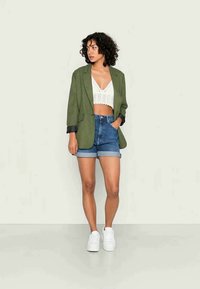 Blazer xadrez verde sobre um top de crochê branco, combinado com calções de denim azuis e ténis brancos. O conjunto apresenta texturas variadas.