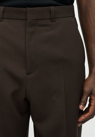 Κοντινή λήψη ενός ατόμου που φοράει σκούρα καφέ γραμμένα παντελόνια, με ορατές θηλιές ζώνης και μέρος του δεξιού χεριού δίπλα στο παντελόνι.