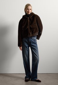 Chaqueta corta de pelaje sintético marrón con capucha, dos bolsillos frontales y dobladillo elástico. Combinada con unos jeans de mezclilla de pierna ancha en azul claro.