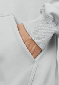 Tejido de sudadera gris claro con un puño acanalado. Una mano reposa en un bolsillo frontal, mostrando una ligera textura y detalles de costura.