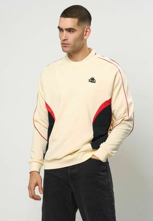 Férfi, aki bézs színű pulóvert visel piros és fekete panelekből, logó a mellkasán, fekete farmert, egyik kezével a zsebében áll, oldalra néz.