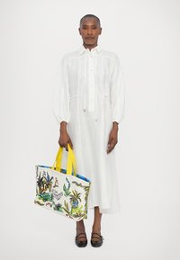 Abito bianco in lino con maniche a sbuffo e fiocco davanti. Tiene una grande borsa tote con stampe botaniche colorate e spalline gialle. Scarpe nere.