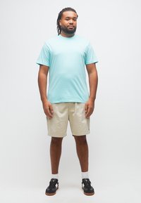 Άνδρας με ντραντλοκς που φοράει ανοιχτό μπλε μπλουζάκι, μπεζ σορτς, άσπρες κάλτσες και μαύρα αθλητικά παπούτσια, στέκεται μπροστά σε απλό λευκό φόντο.