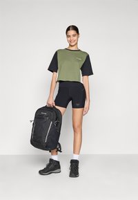 T-shirt cropped a maniche corte color verde oliva e nero, pantaloncini neri, robusti scarponi da trekking neri e uno zaino nero con accenti grigi.