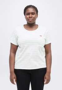 Ενήλικη γυναίκα με πλεξούδες στα μαλλιά, φοράει χρυσούς κρίκους στα αυτιά και ένα ανοιχτό πράσινο ριγέ μπλουζάκι Levi's με μαύρο παντελόνι, στέκεται μπροστά σε λευκό φόντο.