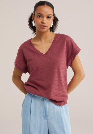 Femeie cu păr creț, purtând cercei albi ovali, un top texturat bordo cu decolteu în V și pantaloni albastru deschis, stând cu mâinile la spate.