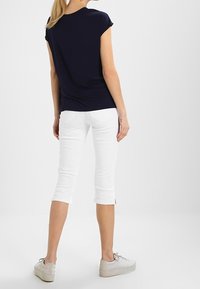 Kvinna klädd i en marinblå kortärmad topp, vita capribyxor och vita sneakers, stående med ryggen mot kameran på en helt vit bakgrund.