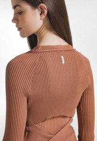 Top marrone a maniche lunghe con design aderente e costine. Presenta un taglio a strati e un piccolo etichetta rosa sul retro vicino al collo.