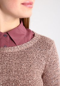 Strickpullover in sanftem Hellbraun mit strukturierter Oberfläche und dezenten Pailletten, darüber ein kastanienbraunes Hemd mit Knöpfen und Kragen getragen.