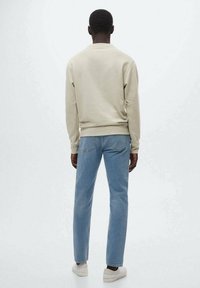 Uomo in piedi, di spalle, che indossa una felpa beige chiaro, jeans blu e sneakers bianche su uno sfondo bianco neutro.