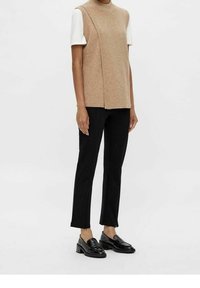 Personne portant un haut beige sans manches en maille par-dessus une chemise blanche à manches courtes, un pantalon noir ajusté et des mocassins à talons noirs, debout.