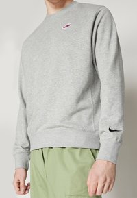 Man som bär en ljusgrå långärmad sweatshirt med en liten röd sko-patch på bröstet och en svart Nike-logga på ärmen, kombinerat med ljusgröna byxor.