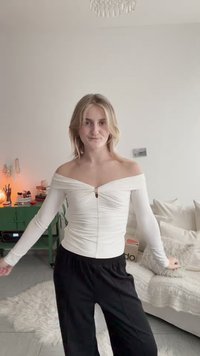 Jeune femme aux cheveux blonds portant un haut blanc à épaules dénudées et un pantalon noir, debout dans un salon chaleureux et bien éclairé avec des coussins et un meuble vert.