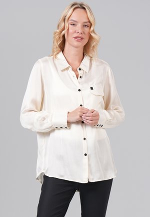 Blouse crème à manches longues avec des boutons noirs, détail de poche et fentes sur les côtés, associée à un pantalon noir. Texture de tissu lisse.