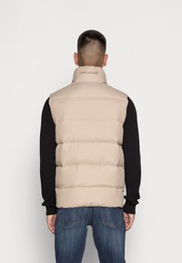 Beige pufferväst med hög krage, horisontell sömnad och logotyp på baksidan, bärs över en svart långärmad skjorta och jeans.