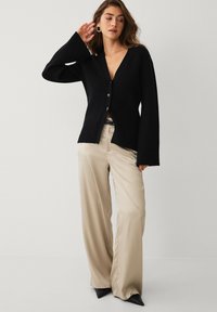 Zwarte cardigan met lange mouwen, V-hals en knoopsluiting, gecombineerd met beige wijde pijpen broek en zwarte schoenen met puntige neus.