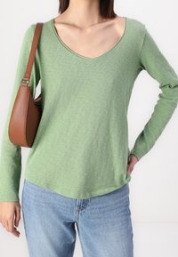 Maglietta verde a maniche lunghe con scollatura a V, caratterizzata da una texture morbida. Abbinata a jeans azzurri chiari e a una borsa marrone con un design curvoso.