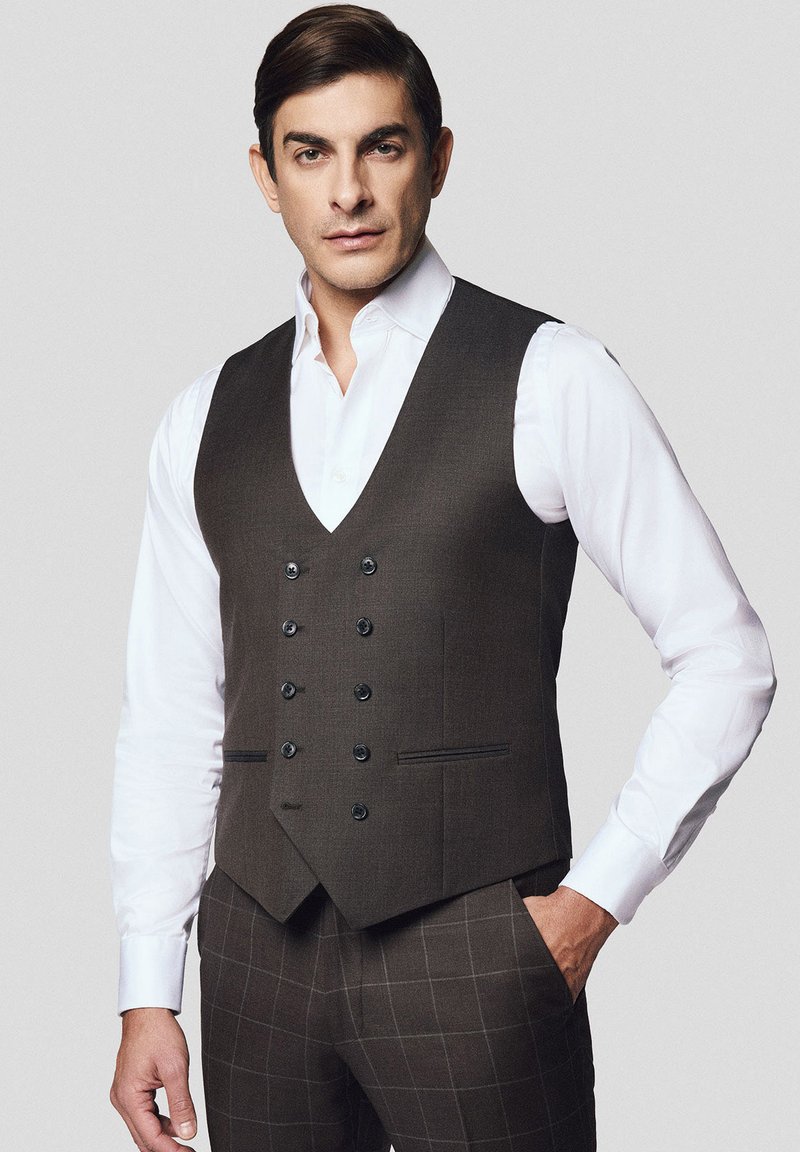 Brązowa, dopasowana kamizelka z dekoltem w kształcie litery V, z podwójnymi guzikami, kieszeniami bocznymi i gładką teksturą, noszona na białej koszuli.