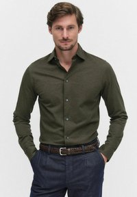 Chemise à manches longues vert olive avec un col boutonné et des boutons bleu marine, assortie à un pantalon bleu foncé et une ceinture tressée marron.