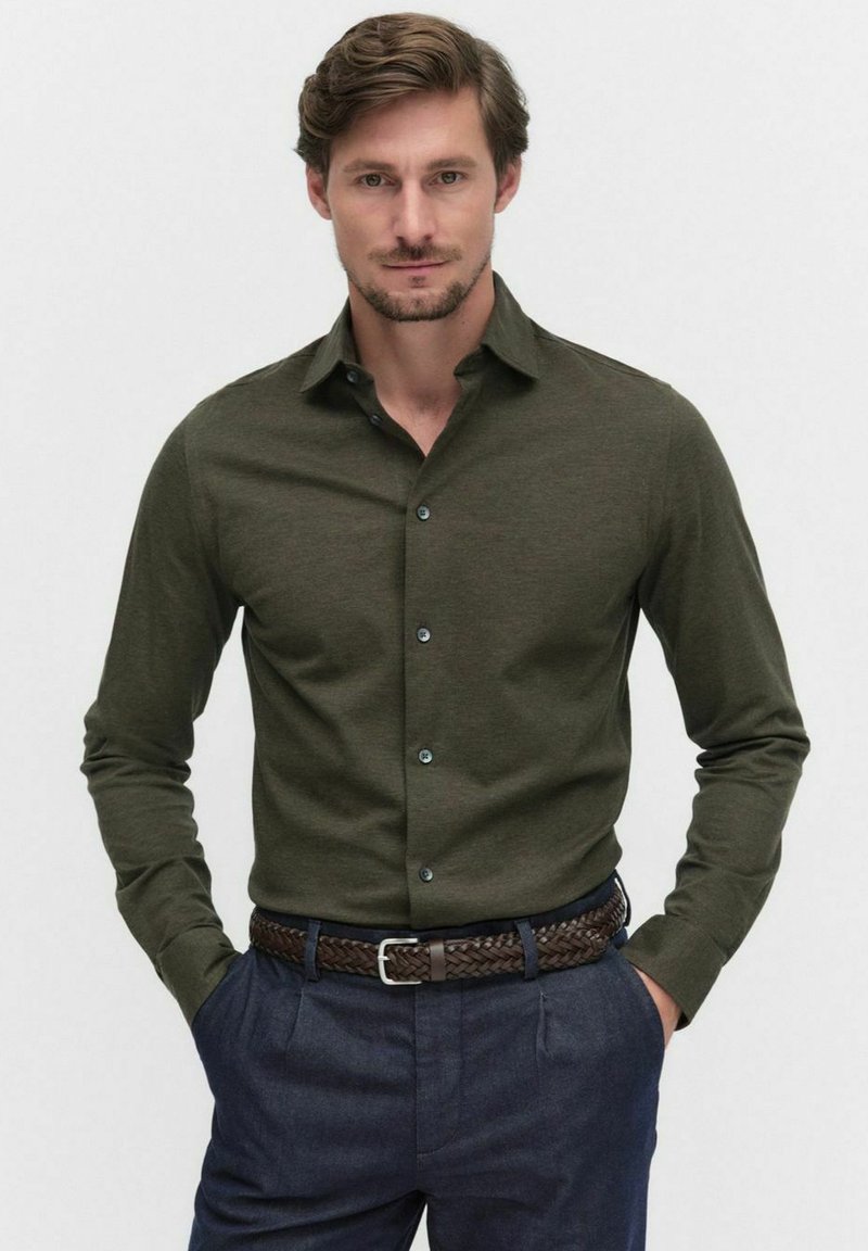 Chemise à manches longues vert olive avec un col boutonné et des boutons bleu marine, assortie à un pantalon bleu foncé et une ceinture tressée marron.