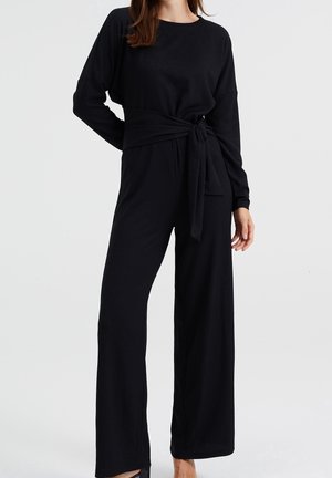 Schwarzer Jumpsuit aus strukturiertem Stoff, mit lockerem Schnitt, langen Ärmeln und einem Taillendetail zum Binden. Die Hose hat einen weitem Bein.