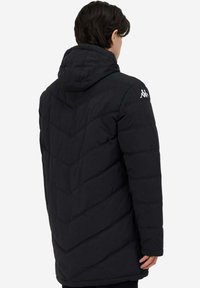 Giacca a vento nera con cappuccio, caratterizzata da trapuntatura a V, maniche lunghe e un logo sulla spalla sinistra. Texture liscia e taglio fino al ginocchio.