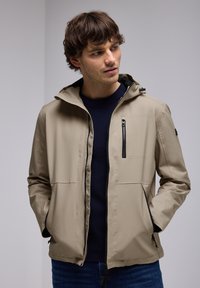 Veste légère beige avec capuche, fermeture éclair à l'avant et poches latérales. Comprend une poche poitrine zippée et des accents noirs sur la fermeture éclair et la doublure.