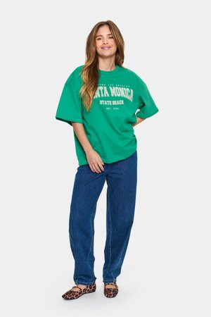 T-shirt oversize vert avec "SANTA MONICA STATE BEACH EST. 1989" imprimé en blanc, associé à un jean large bleu et des ballerines à imprimé léopard.
