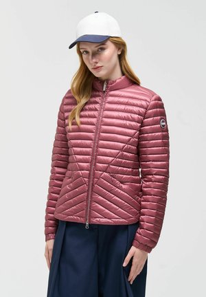 Vrouw draagt een mauve gewatteerde jas met diagonale stiksels, navy broek en een wit-blauwe baseballpet, staand tegen een grijze achtergrond.